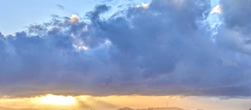 Dark gray-blue, puffy cloud across a wide slice of the sky, highlights from the setting sun touch the top of the clouds, and beam out beneath the cloud in golden light.